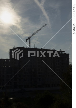 Tower cranes used for installing roof over unfinished concrete building, concept of vertical urbanization. Heavy construction machinery arms extended over high rise building against blue cloudy sky Tower cranes used for installing roof over unfinished concrete building, concept of vertical urbanization. Heavy construction machinery arms extended over high rise building against blue cloudy sky 135817902