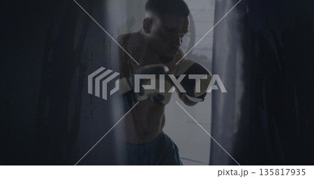 Striking shirtless boxer hitting heavy bag in dim gym room, wearing boxing gloves and blue shorts Striking shirtless boxer hitting heavy bag in dim gym room, wearing boxing gloves and blue shorts 135817935