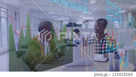 Gesturing bomber-jacketed man, listening checkered-blouse woman discussing data on monitors at desk 135818164