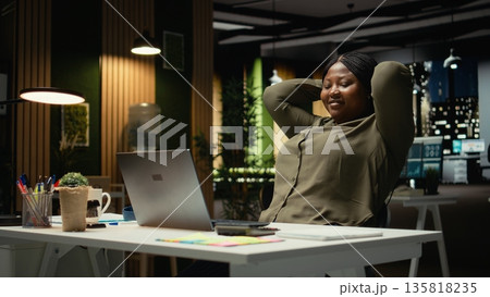 African american woman reclining in a chair in a relaxed pose at midnight, daydreaming during work break. Fulfilled and self assured, reflecting on career success and achievements. Camera A. African american woman reclining in a chair in a relaxed pose at midnight, daydreaming during work break. Fulfilled and self assured, reflecting on career success and achievements. Camera A. 135818235