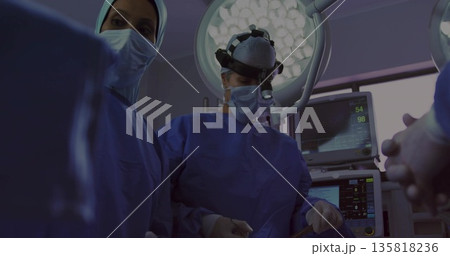 Operating female surgeons in blue gowns working on patient in OR, with overhead light, headlamp Operating female surgeons in blue gowns working on patient in OR, with overhead light, headlamp 135818236