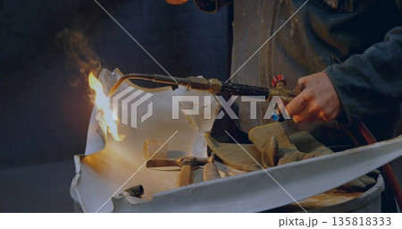 Shaping metal panel metalworker wearing jacket using blowtorch on drum in shop, with hammers mallet 135818333