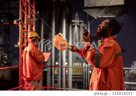 African american engineer exchanges insights on walkie talkie from drilling platform, coordinating fuel and natural petroleum extraction on offshore oil rig. Efficient fossil fuels production. 135818551