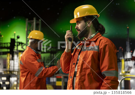 Offshore rig employee using walkie talkie to coordinate logistics at night under aurora borealis. Drilling barge radio operator supervising deck machinery, with green northern lights in background Offshore rig employee using walkie talkie to coordinate logistics at night under aurora borealis. Drilling barge radio operator supervising deck machinery, with green northern lights in background 135818566