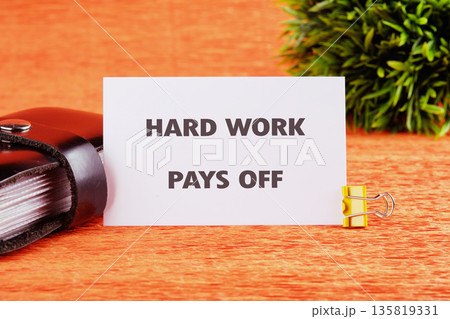 A white card bearing the phrase HARD WORK PAYS OFF rests on an orange surface, held by a yellow binder clip 135819331