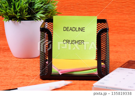 A bright green sticky note with DEADLINE CRUSHER sits in a black mesh organizer. It is on an orange wooden desk next to a potted green plant and a white pencil, showing office inspiration A bright green sticky note with DEADLINE CRUSHER sits in a black mesh organizer. It is on an orange wooden desk next to a potted green plant and a white pencil, showing office inspiration 135819338
