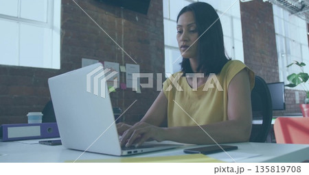Typing mature Asian woman using silver laptop at modern open-plan office desk, with coffee cup Typing mature Asian woman using silver laptop at modern open-plan office desk, with coffee cup 135819708