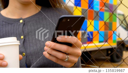 Holding smartphone and coffee cup, woman leaning by chain link fence, with colorful delivery truck Holding smartphone and coffee cup, woman leaning by chain link fence, with colorful delivery truck 135819987