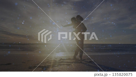 Giving child piggyback ride on sandy beach at sunset, silhouetted parent showing lens flare overlay 135820075