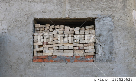 Used gray bricks stacked in a window opening in the center of a high concrete wall, abandoned unfinished dilapidated building with ruin details, old warehouse window with a pile of bricks Used gray bricks stacked in a window opening in the center of a high concrete wall, abandoned unfinished dilapidated building with ruin details, old warehouse window with a pile of bricks 135820623
