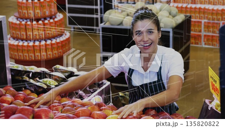 Organizing clerk in green-striped apron leaning over red apples at market produce, with juice trays 135820822