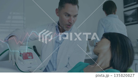Demonstrating male doctor explaining defibrillator use in clinic, with exam chair Demonstrating male doctor explaining defibrillator use in clinic, with exam chair 135820827