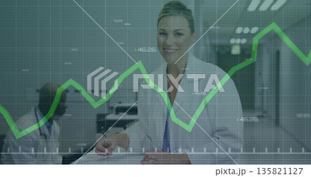 Smiling doctor wearing lab coat writing on clipboard in clinic corridor with green graph overlay 135821127