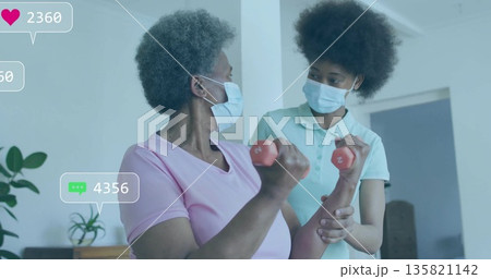 Guiding therapist supporting masked senior woman lifting red dumbbells at home, with overlay icons 135821142