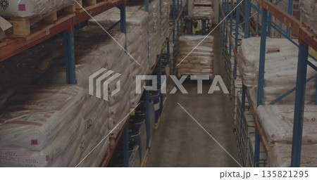 Driving forklift carrying pallet of wrapped sacks down warehouse aisle, with racks and drums Driving forklift carrying pallet of wrapped sacks down warehouse aisle, with racks and drums 135821295