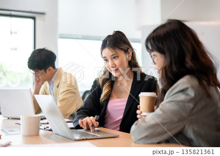 Smiling asian businesswoman showing data in laptop to coworker while sits at wooden table in office. 135821361