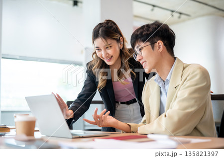Smiling asian businesswoman looking at laptop talking with intern coworker at wooden table in office 135821397