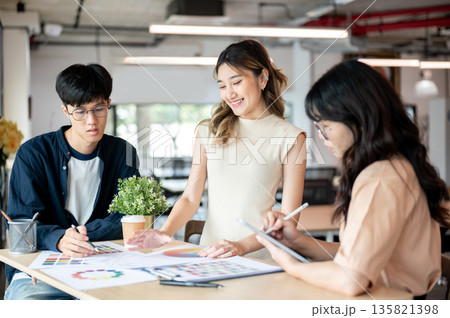Smiling asian woman designer looking at color chart and talking with coworkers at table in office 135821398