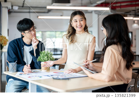 Smiling asian woman designer talking with coworkers about color chart at wooden table in office. 135821399