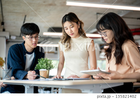 Pretty asian woman designer talking with coworkers over documents at wooden table in office. Pretty asian woman designer talking with coworkers over documents at wooden table in office. 135821402