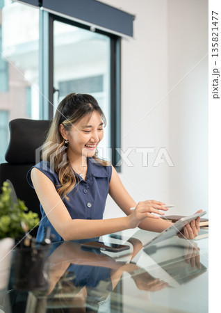 Asian woman office worker holding stylus and looking at tablet while sits at glass table in office. 135821477