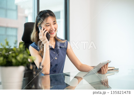 Asian woman office worker holding tablet and talking on phone while sitting at glass table in office 135821478