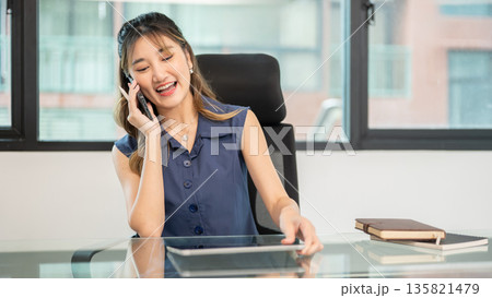 Asian woman office worker putting down tablet as talking on phone while sit at glass table in office Asian woman office worker putting down tablet as talking on phone while sit at glass table in office 135821479