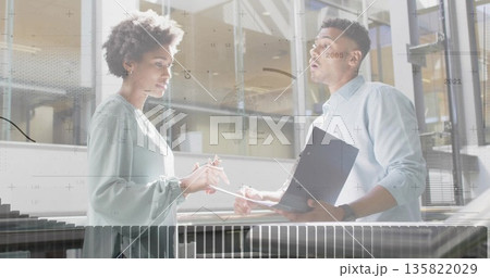Discussing data two colleagues reviewing reports in modern atrium, with folder pen railing glass Discussing data two colleagues reviewing reports in modern atrium, with folder pen railing glass 135822029