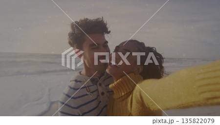 Smiling couple in yellow turtleneck and striped sweater taking selfie on beach, with smartphone 135822079