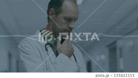 Standing mature doctor contemplating in hospital hallway, in white coat with stethoscope and badge Standing mature doctor contemplating in hospital hallway, in white coat with stethoscope and badge 135822153