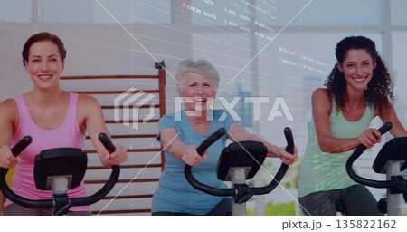 Pedaling three women wearing colorful tops at gym with bikes, wall bars and window 135822162