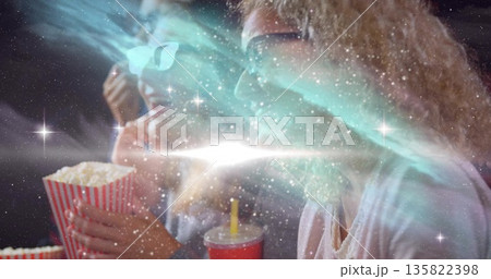 Holding popcorn, red cup, two women wearing 3D glasses watching movie under cosmic light at theater 135822398