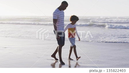 Father and son walking barefoot along wet sandy shoreline under bright sun, with ocean waves 135822403