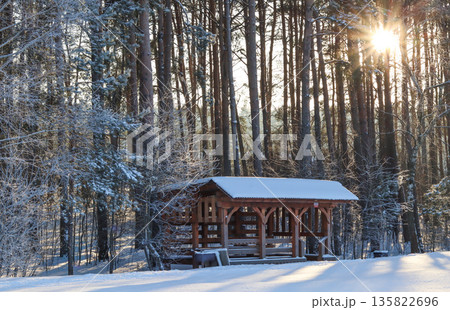 Wooden gazebo standing in a snowy winter forest capturing warm sunlight through pine trees on a cold morning Wooden gazebo standing in a snowy winter forest capturing warm sunlight through pine trees on a cold morning 135822696