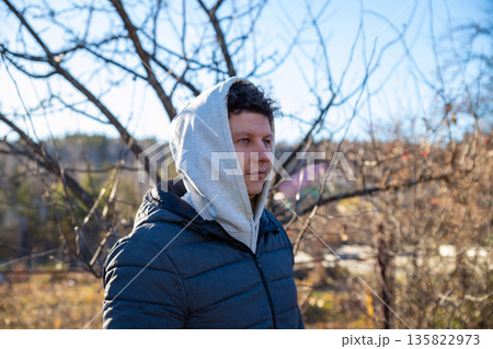 young Caucasian man with curly hair and hood on his head in autumn garden on sunny day. Landowner, farmer, organic farm worker, weekend hiking, healthy lifestyle, race walking. waist up portrait 135822973
