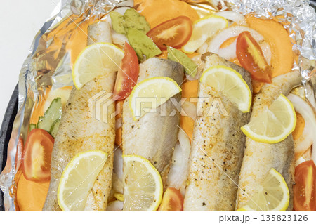 Prerape raw spiced hake for a bake in the oven with lemon, tomatoes, carrots, and onions in foil. Delicious fish for lunch or dinner, perfect for a diet. Tasty and fresh  hake fish for bake with 135823126