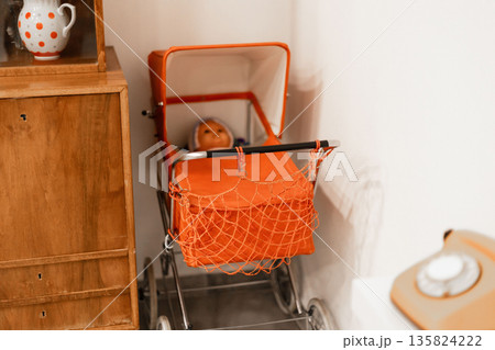 A charming vintage doll stroller, featuring a bright orange color, holds a doll inside. The setting reflects a cozy and nostalgic atmosphere, with wooden furniture and decor 135824222