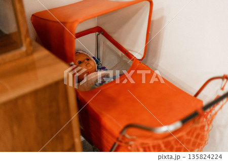 A vintage doll lies quietly in a bright orange stroller, surrounded by a warm and inviting room setting, highlighting a nostalgic atmosphere A vintage doll lies quietly in a bright orange stroller, surrounded by a warm and inviting room setting, highlighting a nostalgic atmosphere 135824224
