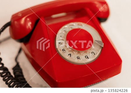 A bright red rotary phone sits on a plain white background, showcasing its retro design and circular dial. This vintage telephone evokes nostalgia for past communication methods 135824226