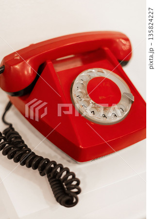 A vintage red rotary telephone is positioned on a smooth white surface. The warm light highlights its glossy finish and the coiled cord, evoking nostalgia A vintage red rotary telephone is positioned on a smooth white surface. The warm light highlights its glossy finish and the coiled cord, evoking nostalgia 135824227