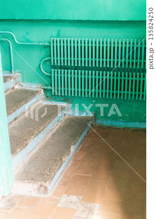 The interior of an old house reveals worn-out steps leading to a bright green wall. A rusty radiator adds to the neglected atmosphere. Dust and marks indicate years of disrepair 135824250