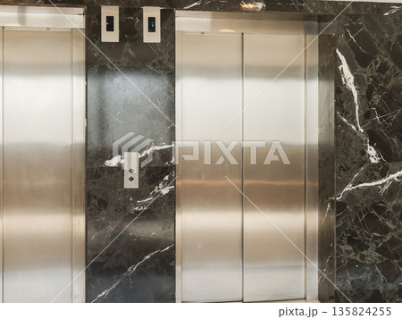 Two elevator doors stand in a building with dark marble walls. The setting shows a clean and modern environment for accessing different floors of the structure Two elevator doors stand in a building with dark marble walls. The setting shows a clean and modern environment for accessing different floors of the structure 135824255