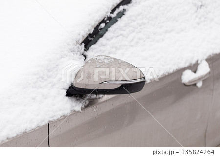 A car parked on a street is covered in snow during winter. The side mirror is almost completely buried beneath the snow. The scene shows the aftermath of a snowstorm 135824264