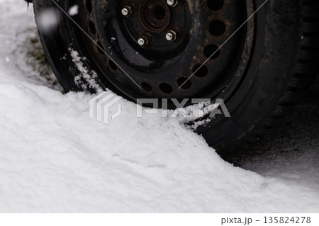 A car tire rests on the ground with snow surrounding it. The tire appears to be stuck in the snow while flakes fall nearby. The scene shows a typical winter day A car tire rests on the ground with snow surrounding it. The tire appears to be stuck in the snow while flakes fall nearby. The scene shows a typical winter day 135824278