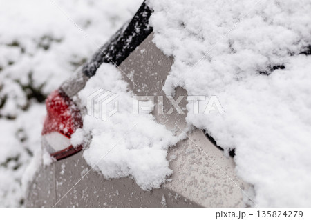 Snow accumulates on the rear of a parked car in a neighborhood. Flakes cover the surface, showing a typical winter scene with overcast skies 135824279
