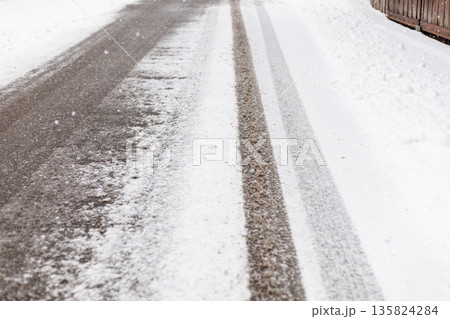 A road is covered with snow and shows clear tire tracks. The surrounding area has unplowed snow, indicating recent snowfall. It is winter A road is covered with snow and shows clear tire tracks. The surrounding area has unplowed snow, indicating recent snowfall. It is winter 135824284