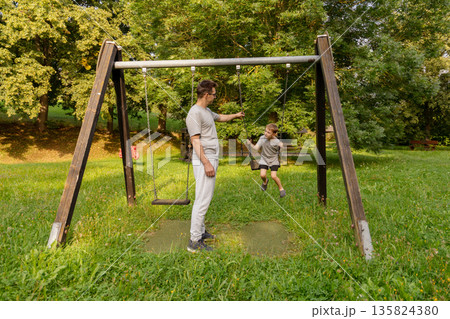 A father engages with his child at a playground, encouraging them while swinging. They share joyful laughter amidst greenery, highlighting the bond they have in this outdoor setting A father engages with his child at a playground, encouraging them while swinging. They share joyful laughter amidst greenery, highlighting the bond they have in this outdoor setting 135824380