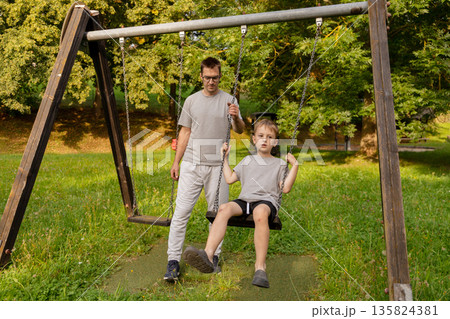 A father and his child enjoy playful moments on a swing set in a vibrant playground. The sun filters through the trees, creating a warm, joyful atmosphere filled with laughter 135824381