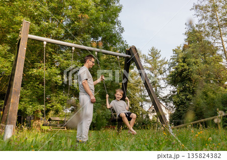 A father pushes his smiling child on a swing, both wearing matching gray outfits. They share joyful laughs in a beautiful park setting, filled with trees and greenery A father pushes his smiling child on a swing, both wearing matching gray outfits. They share joyful laughs in a beautiful park setting, filled with trees and greenery 135824382
