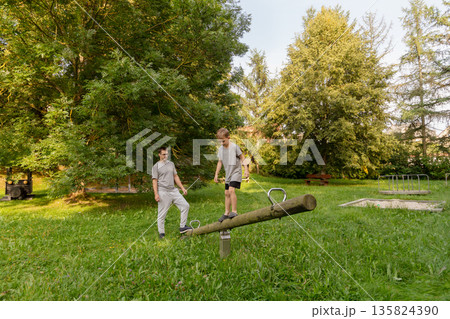 A father watches with joy as his child balances on a wooden seesaw at a playground. They are surrounded by lush greenery, creating a peaceful and playful outdoor atmosphere A father watches with joy as his child balances on a wooden seesaw at a playground. They are surrounded by lush greenery, creating a peaceful and playful outdoor atmosphere 135824390